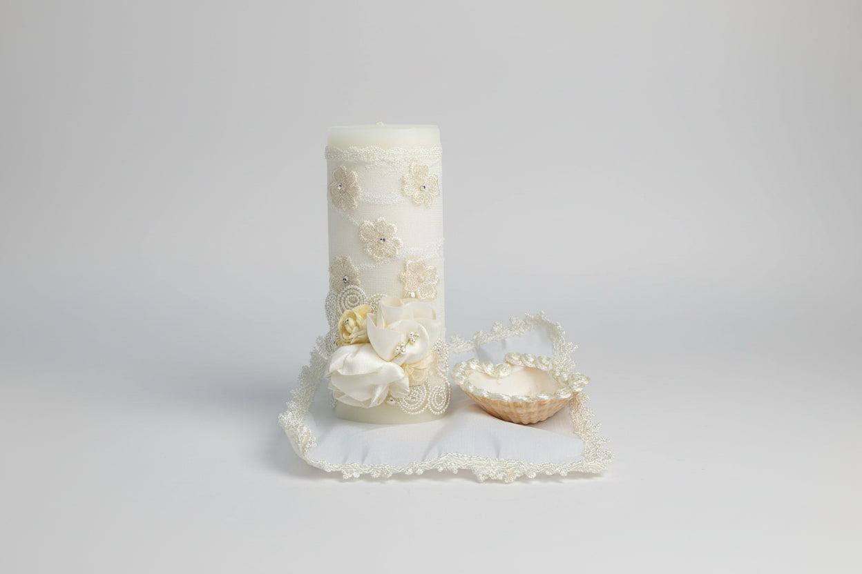 A decorated ivory candle with a shell hankerchief and a small white cloth with a floral pattern, displayed against a backdrop of pink and white flowers.