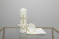 A three-piece candle set for boys, featuring a decorated candle with a cross design, a white handkerchief, and a shell, displayed in front of a floral backdrop.
