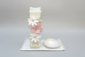 A decorated ivory candle set against a floral background, part of a 3 piece girl baptism candle set, with a shell hankerchief and a small container.