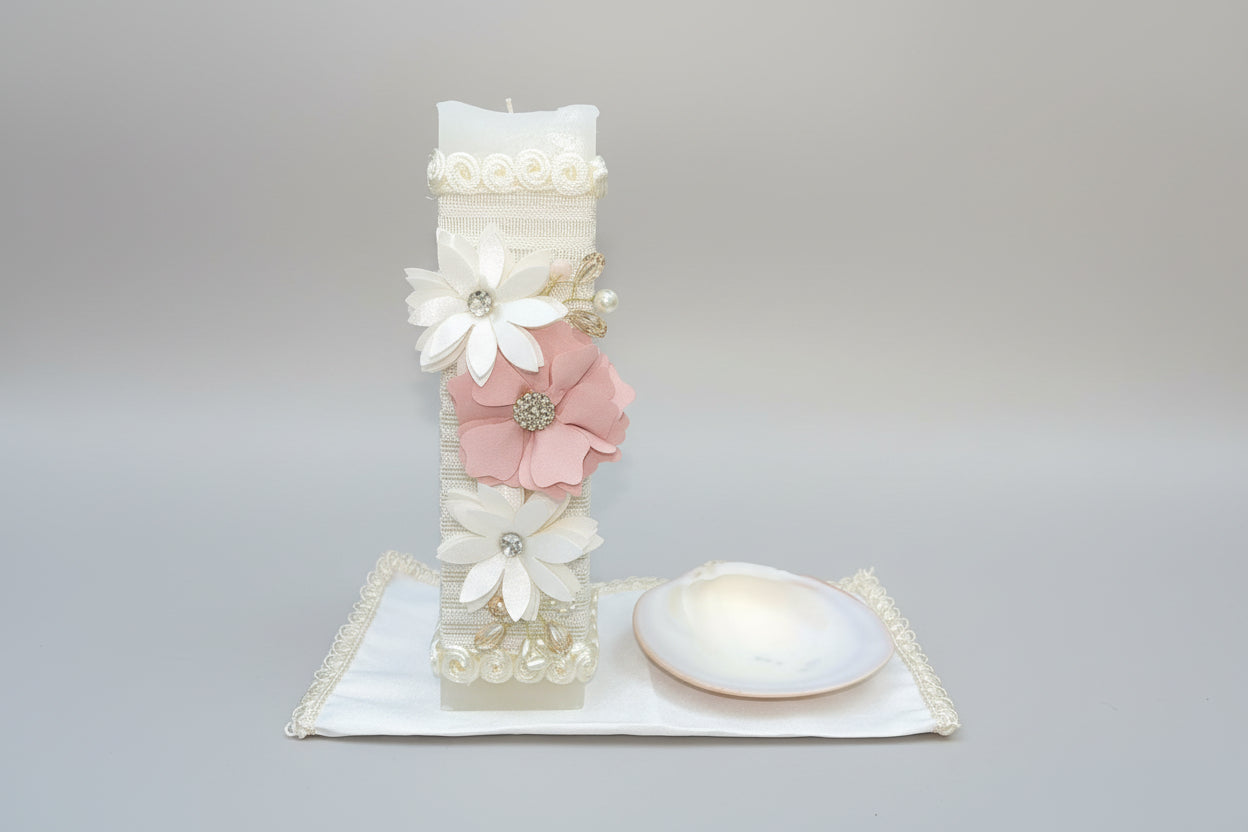 A decorated ivory candle set against a floral background, part of a 3 piece girl baptism candle set, with a shell hankerchief and a small container.