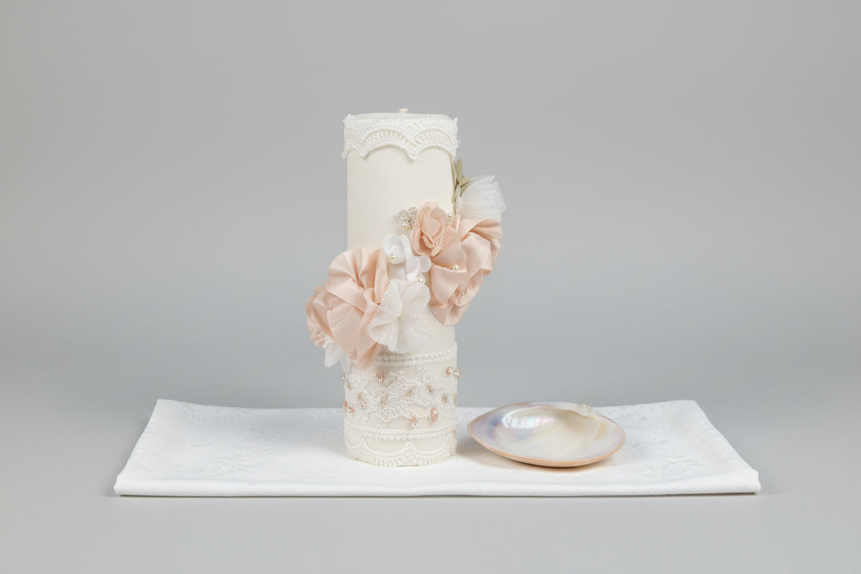 An ivory colored candle set with a decorated candle, shell hankerchief, and a small pink candle, displayed in front of a floral backdrop.