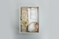 A 3 piece girl baptism candle set displayed in an open box, showing a decorated candle with a floral pattern, a natural shell, and a lace hankerchief with gold accents.