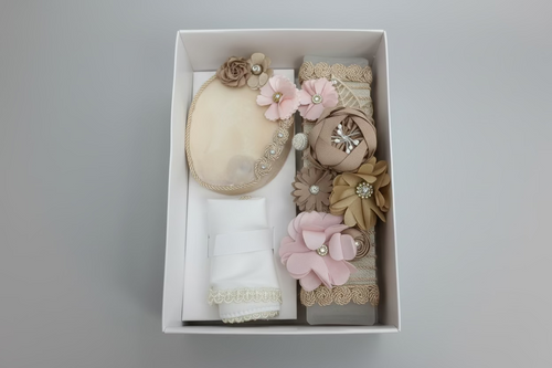 A 3 piece girl baptism candle set displayed in an open box, including a decorated candle with pink and brown flowers, a natural shell, and a hankerchief with champagne style decorations.